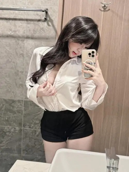 Trước gương phòng kín, cô gái che mặt bằng điện thoại, áo sơ mi trắng phối short đen tạo dáng nghiêng với mái tóc rủ nhẹ