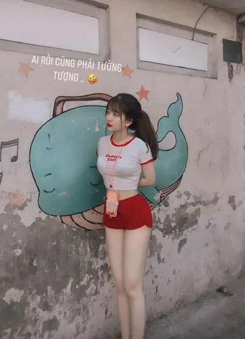 Bức tường vẽ hình cá xanh tạo nền vui mắt, cô gái áo trắng quần short đỏ đứng nghiêng với nụ cười rất nhẹ