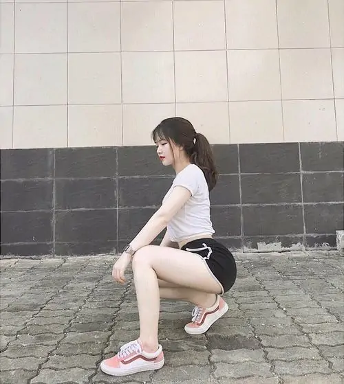 Cô gái áo trắng và short đen ngồi xổm bên mảng tường gạch, đôi giày hồng tạo điểm nhấn trẻ trung cho khung hình