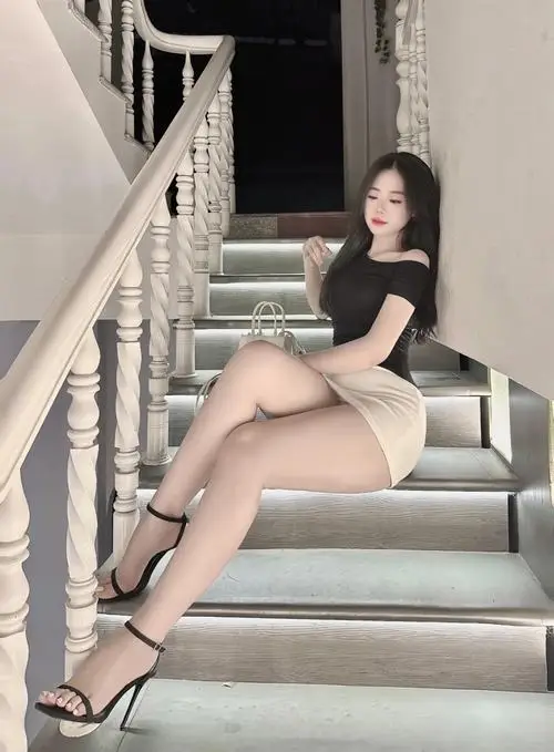 Cầu thang sang trọng làm nền cho cô gái mặc váy đen ngắn, đôi chân dài và dáng ngồi nghiêng tạo vẻ thanh lịch