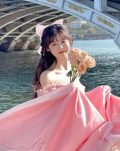 Cô gái ngồi bên hồ nước trong xanh, váy hồng xòe rộng bắt sáng khiến dáng pose trông dịu dàng và bay bổng