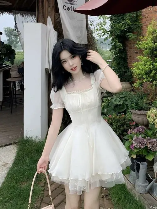 Dưới tán cây trong vườn, cô gái mặc váy trắng xòe cầm túi nhỏ, tay vuốt tóc và dáng đứng nhẹ nhàng