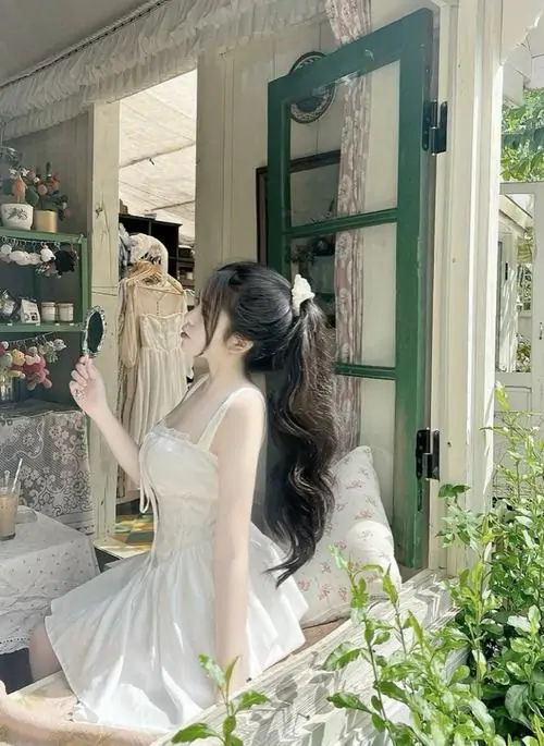 Gái xinh mặc váy trắng ngắn đứng trong quán sáng màu, mái tóc dài buộc nơ và dáng nghiêng nhìn rất thanh thoát
