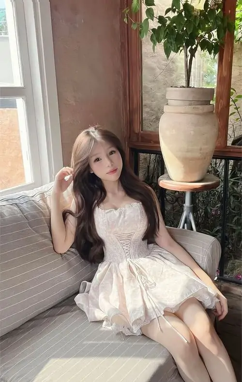 Bên cửa sổ phủ rèm hồng, cô gái váy trắng ngồi co chân, mái tóc nâu dài và ánh nhìn nhẹ nhàng