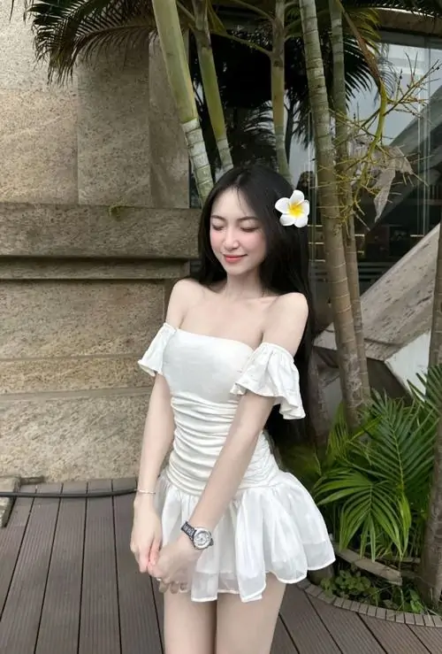 Cạnh cầu thang gỗ, ảnh người đẹp diện váy trắng ngắn nổi bật với vai trần, nụ cười tươi và dáng đứng tự tin
