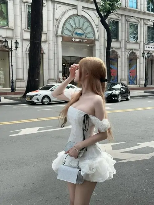 Trên phố rộng ban ngày, cô nàng váy trắng xếp tầng chụp nghiêng từ sau, túi nhỏ và tóc nâu tạo nét nhẹ