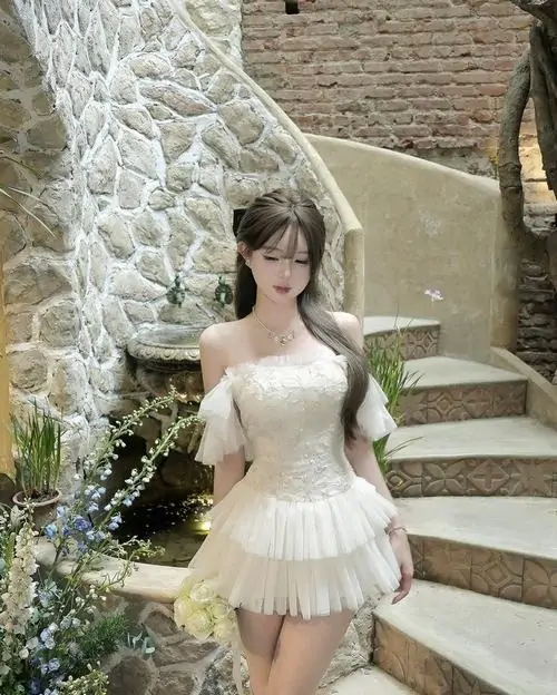 Cô gái váy trắng xòe đứng cạnh cầu thang đá, tay cầm hoa nhỏ, ánh mắt nhìn xa tạo cảm giác mềm mại