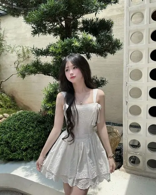 Gái xinh mặc váy trắng ngắn đứng trong khu vườn xanh, váy hai dây bồng nhẹ và mái tóc dài phủ trước ngực