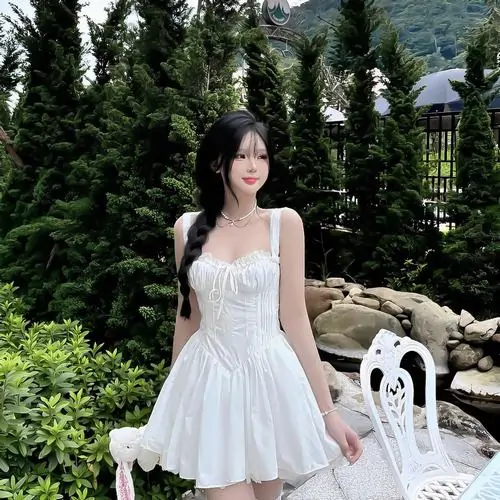 Giữa nền cây tối màu, ảnh cô gái mặc đầm trắng dáng ngắn nổi bật với nụ cười tươi và dáng đứng ngay ngắn