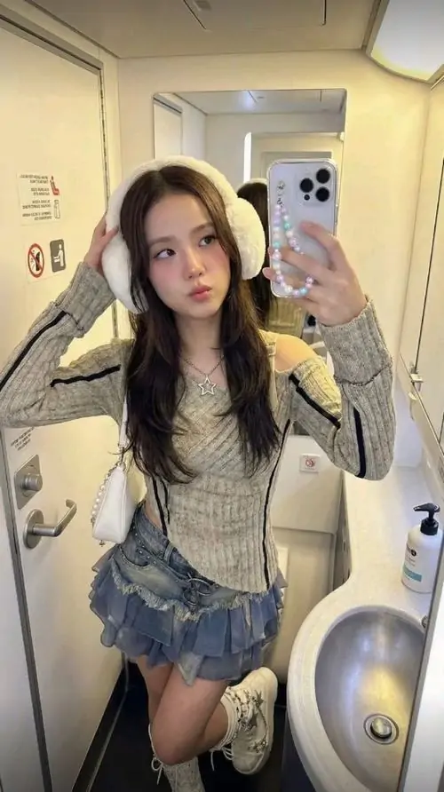 Hành lang khách sạn sáng vàng làm nổi bật cô gái tóc dài mặc áo trắng, váy denim ngắn và biểu cảm tự tin trước gương