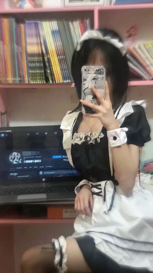 Ảnh hầu gái trước kệ sách ghi lại cô gái selfie nhẹ nhàng, tóc tối màu xõa vai và tạp dề trắng nổi bật trên nền đen