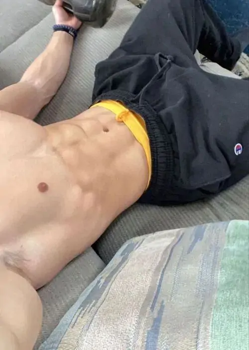 Góc chụp trên giường khá gần, chàng trai mặc quần short tối màu, thân trên săn chắc, tay đặt tự nhiên và ánh sáng phòng ấm