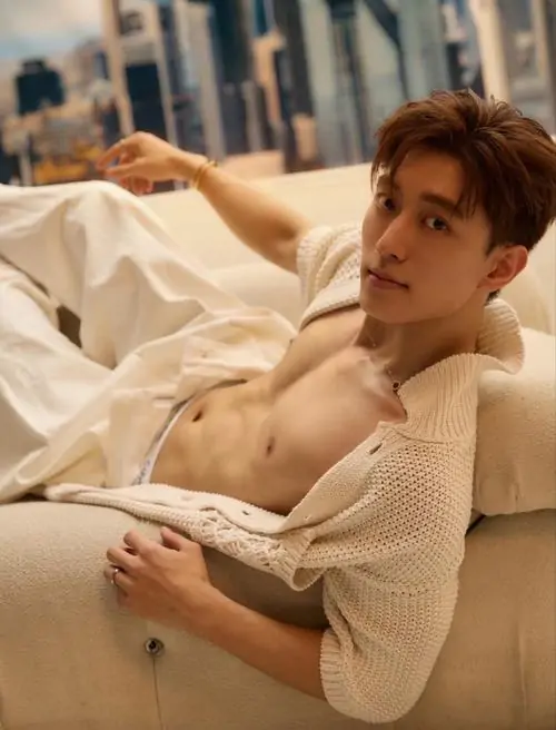 Ánh sáng phòng ngủ ấm áp chiếu lên chàng trai tóc nâu nằm nghiêng, mặc áo len mỏng mở rộng, vẻ mặt thư thái và mềm hơn