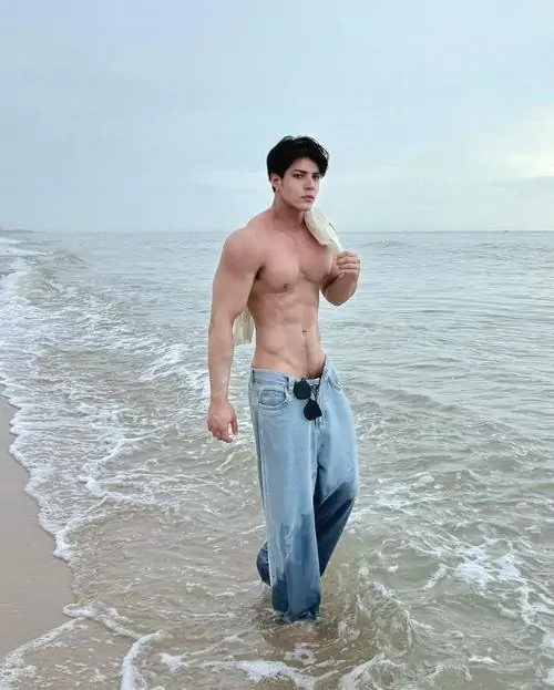 Bãi biển xám nhạt làm nền cho ảnh nam thần thể hình Việt, chàng trai mặc quần jeans, tóc đen gọn và dáng đứng bình tĩnh