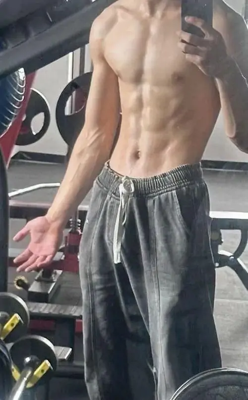Góc chụp trong phòng gym tập trung vào thân trên săn chắc, chàng trai mặc quần thể thao tối màu, tay thả tự nhiên bên máy tập