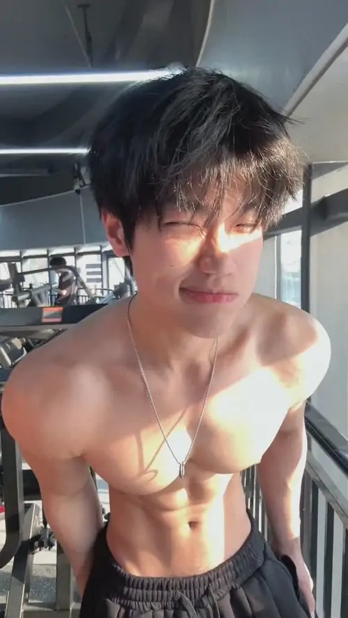 Ánh nắng ngoài trời phủ lên cơ bụng rõ múi, chàng trai tóc đen ướt nhẹ, mặc quần sáng màu và nhìn thẳng khá nghiêm túc