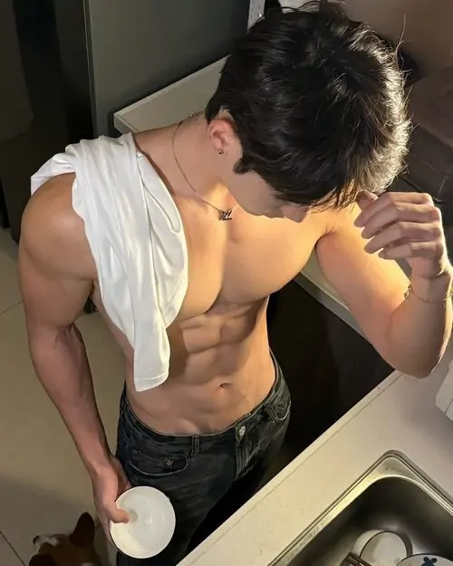 Nam mẫu cúi đầu trước gương, áo trắng vén lên, tay cầm điện thoại và cơ bụng lộ rõ trong không gian phòng tắm sáng