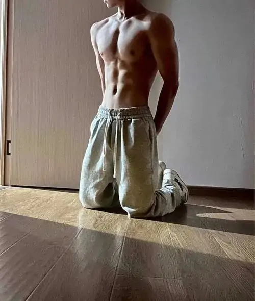 Chàng trai quỳ trên sàn gỗ cạnh cửa sổ, áo kéo cao qua đầu, quần sáng màu và cơ bụng hiện rõ trong ánh sáng tự nhiên