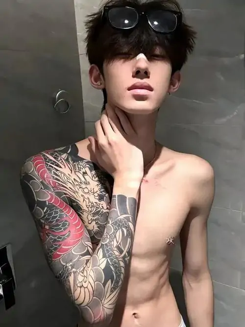 Mái tóc đen phủ trán che nhẹ ánh mắt, ảnh nam thần cơ bắp khoe hình xăm tay và thân trên săn chắc trước nền tường xám