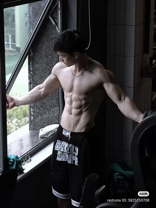 Bên cửa kính lớn, hình thể nam giới sáu múi hiện lên qua dáng đứng nghiêng, quần thể thao đen và ánh sáng tự nhiên hắt mạnh