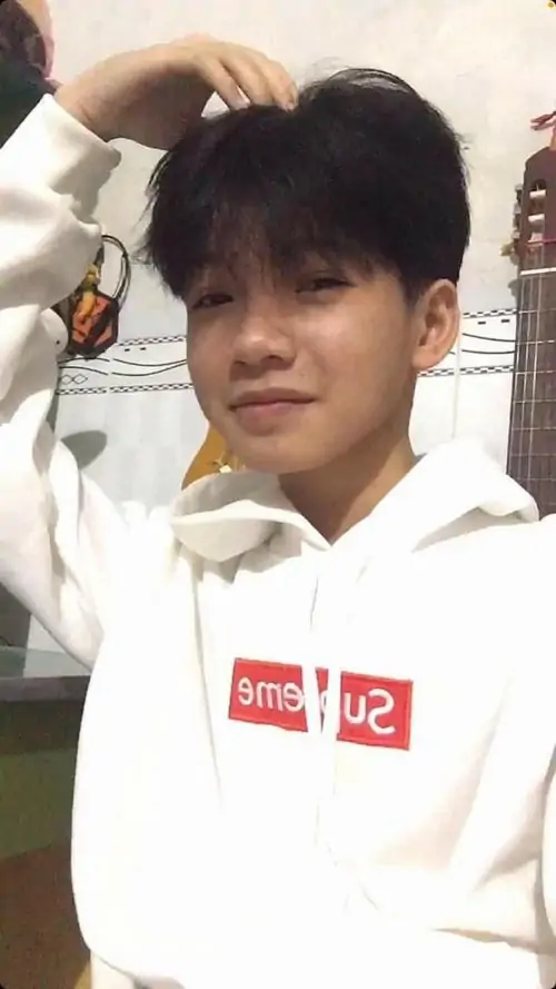 Hoodie trắng in đỏ làm cậu bạn cười nhẹ thêm thân thiện, một tay đưa lên đầu tạo dáng vui vẻ trong không gian trong nhà