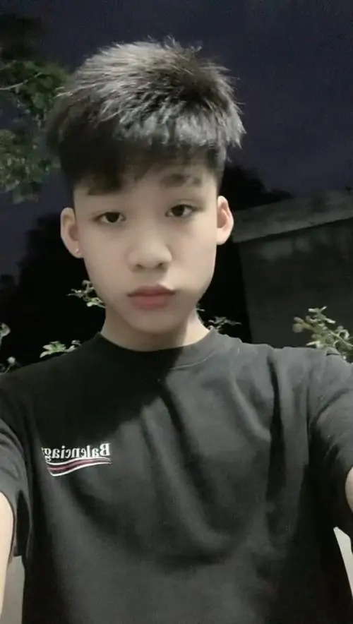 Nền tối ngoài trời làm gương mặt bạn nam áo đen nổi bật hơn, tóc dựng nhẹ và ánh mắt nhìn thẳng có vẻ hơi ngại