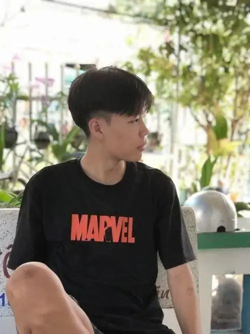 Trai đẹp 2k11 mặc áo thun Marvel ngồi ngoài trời, góc nghiêng dưới bóng cây làm nổi rõ mái tóc dày và dáng vẻ trầm
