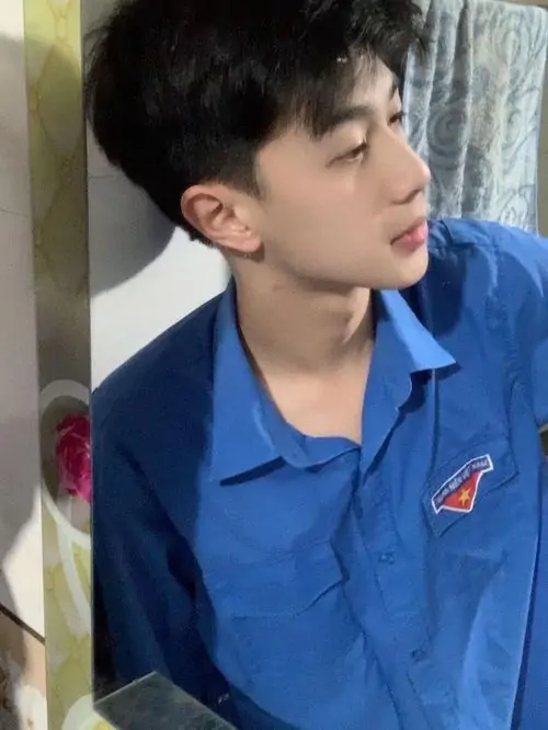 Sơ mi xanh nổi bật dưới ánh sáng trong nhà, cậu bạn quay nghiêng khoe mái tóc cắt gọn và dáng vẻ hơi trầm