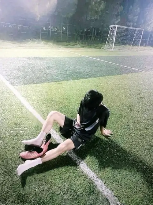 Trên sân cỏ xanh nhạt, K5 mặc đồ đen ngồi duỗi chân cạnh vạch sân, tóc dài che mặt tạo dáng khá lặng