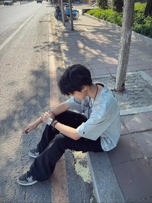 Nắng vỉa hè làm bạn nam áo trắng ngồi bên gốc cây thêm nổi bật, tóc đen rủ và dáng cúi nhìn tay khá tự nhiên