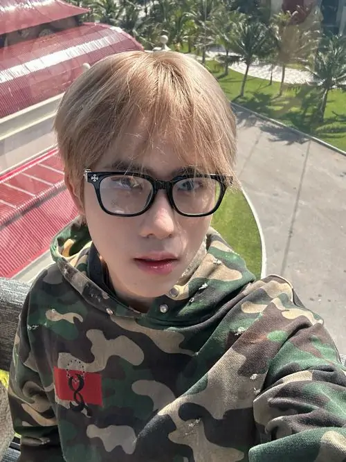 Góc selfie cận mặt của chàng trai tóc vàng đeo kính tròn, áo rằn ri xanh và biểu cảm thẳng máy nhìn rất hiền