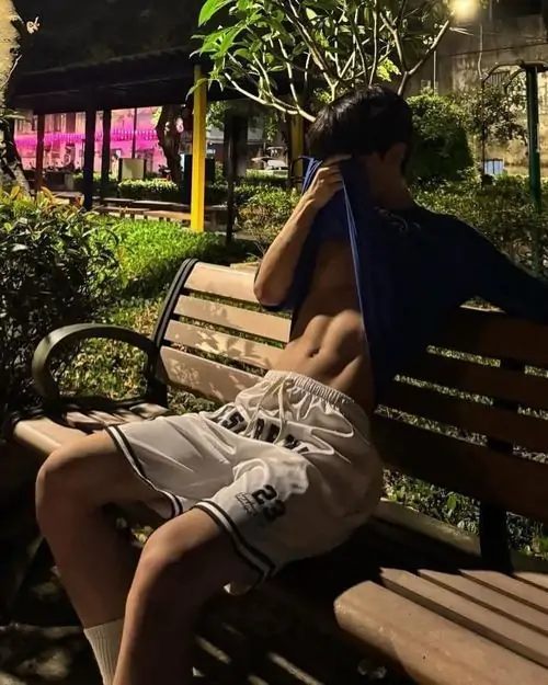 Trên ghế gỗ công viên ban đêm, bạn nam mặc áo sát nách tối màu ngồi nghiêng, ánh đèn vàng khiến khung ảnh ấm hơn