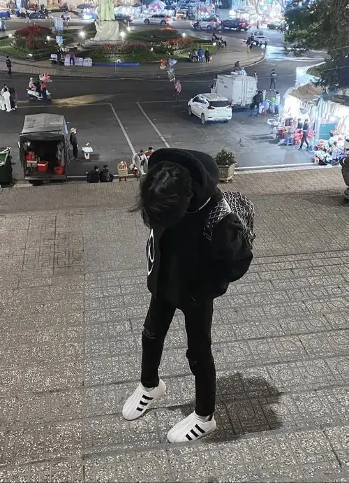 Từ góc chụp cao ngoài phố, chàng trai mặc đồ đen đứng cúi đầu, balo sau lưng và sneaker trắng tạo vẻ gọn gàng