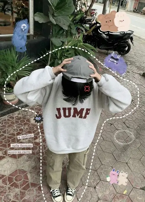 Trên nền gạch sân nhà, bạn nam mặc hoodie trắng chữ đỏ đưa tay chỉnh mũ, quần xanh rêu và sneaker trắng khá trẻ