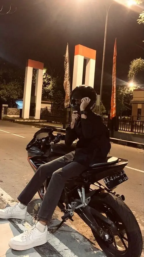 Ánh đèn đêm phủ lên chàng trai ngồi cạnh xe máy, outfit đen và dáng dựa nhẹ tạo cảm giác đường phố rất rõ