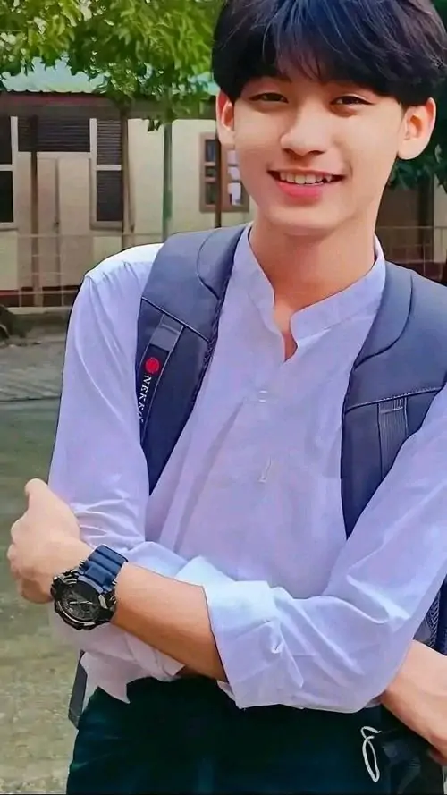 Chàng trai mặc sơ mi trắng đứng cười nhẹ trước sân trường, tóc đen gọn gàng, balo đeo vai tạo cảm giác nam sinh sáng sủa