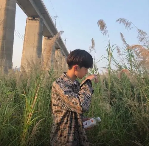 Giữa bãi cỏ lau vàng nhạt, bạn nam áo sơ mi kẻ cúi đầu nhìn điện thoại, mái tóc đen rủ nhẹ tạo nét trầm