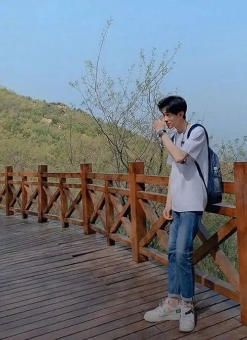Con đường gỗ và bầu trời xanh làm nổi bật bạn nam đeo balo trắng, tóc rủ che trán và dáng đứng nghiêng khá thư sinh