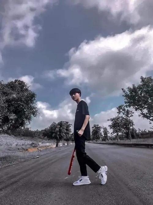 Giữa con đường rộng dưới mây xám, chàng trai áo đen đứng nghiêng, quần trắng và sneaker nổi bật trong tông màu lạnh