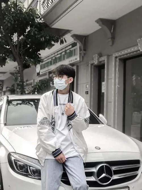 Trước chiếc xe trắng, bạn nam mặc áo khoác sáng màu và khẩu trang, dáng đứng tự tin cùng tóc đen tạo vẻ hiện đại