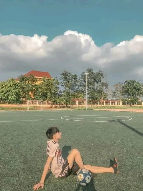 Bầu trời xanh và sân thể thao rộng làm nền cho nam sinh 2008 ngồi thư giãn, áo ba lỗ tối màu tạo vibe năng động