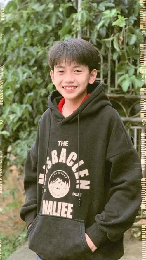 Nụ cười tươi của bạn nam mặc hoodie xanh rêu nổi bật giữa nền cây lá, mái tóc ngắn mềm khiến gương mặt càng dễ mến