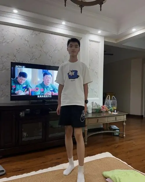 Cậu bé mặc áo trắng đứng trong phòng khách hiện đại với TV phía sau.