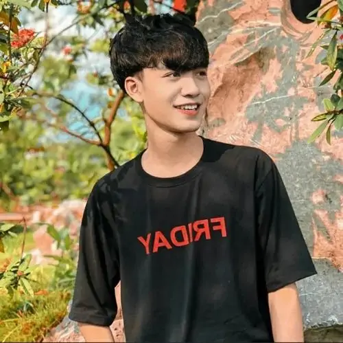 Bạn trai trẻ mặc áo đen in chữ đỏ đang mỉm cười nhẹ giữa nền cây xanh tươi mát.