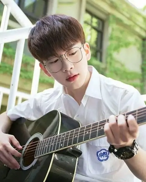 Thiếu niên đeo kính trắng ngồi chơi guitar trong vườn xanh với biểu cảm tập trung.