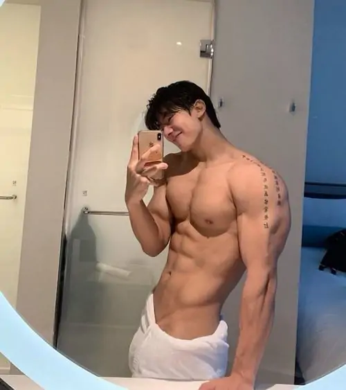 Trong căn phòng sáng, chàng trai tóc đen đứng tạo dáng bên tường, mặc quần trắng, cơ bụng gọn và biểu cảm hơi ngại ngùng