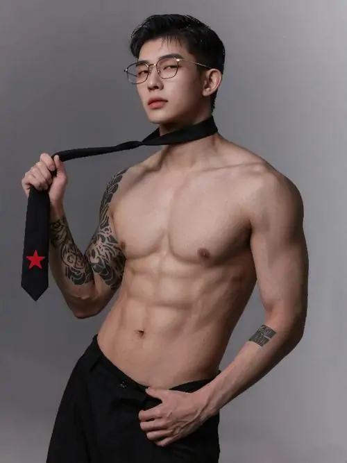 Người mẫu nam mặc quần tối và áo quấn cổ, tạo dáng trong studio xám, cơ ngực dày, bắp tay chắc và gương mặt nghiêm