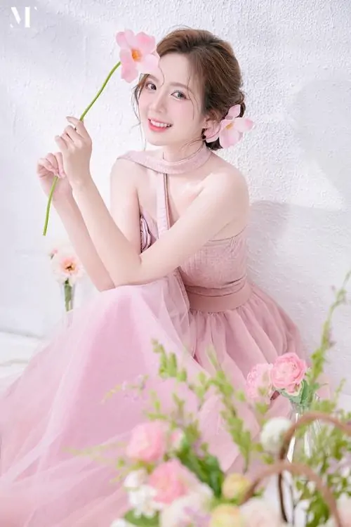 Giữa vườn hoa nhẹ màu, cô bé tóc búi mặc váy hồng tạo dáng cầm chong chóng, gương mặt tươi tắn và đáng yêu