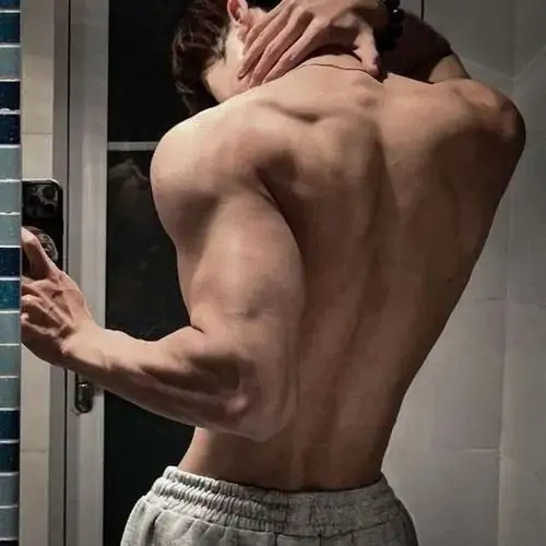 Tông ảnh vàng ấm bao quanh chàng trai tóc sáng mặc áo mở hờ, ngực và bụng hiện nhẹ trong dáng ngồi thư giãn