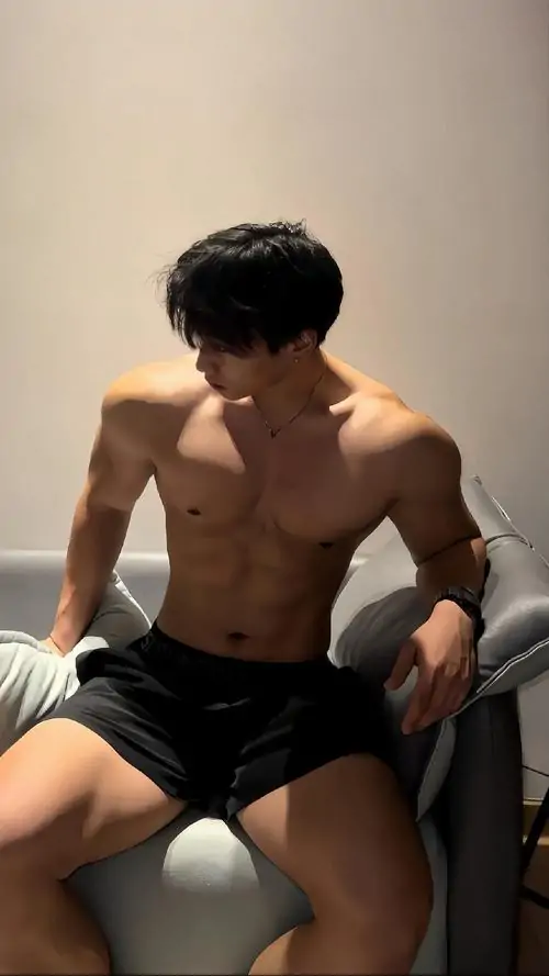 Bên tủ gỗ, chàng trai tóc đen đứng nghiêng với áo ba lỗ hạ thấp, vòng tay đen và cơ vai nổi bật